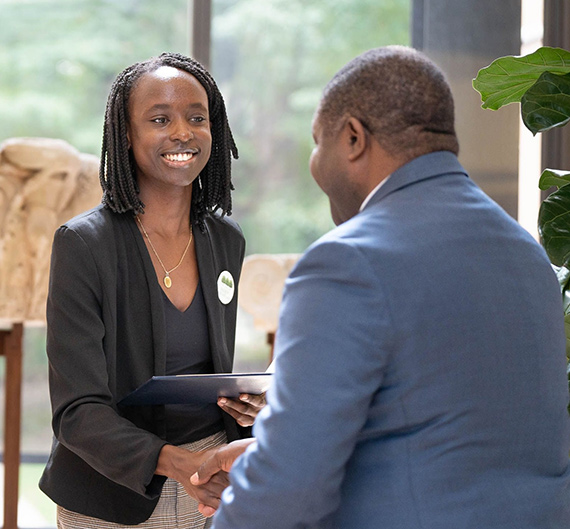 A smiling young black woman shakes hands and presents an award certificate to the ambassador of Zambia