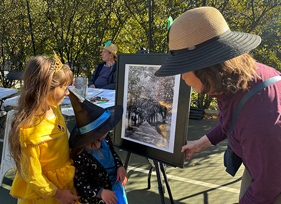 Two children in brightly colored Halloween costumes listen intently while a woman in gardening attire explains the story behind a 1913-era black and white photo of the double row of linden trees shading Washington DC's Massachusetts Ave.