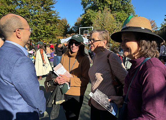 Chinese man in business suit chats with three woman at festive outdoor gathering under bright blue sky and autumn foliage.