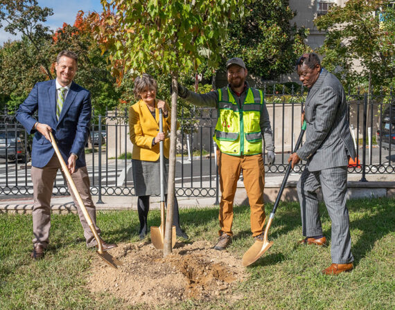 Embassy personnel in business suits and tree keepers in safety vests hold shovels for ceremonial tree planting